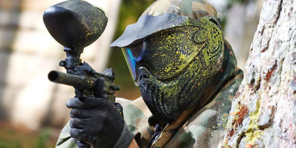 Man hiding behind tree whilst paintballing with yellow paint splattered on his helmet 